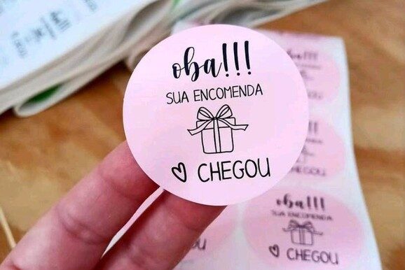
Imagem de adesivo personalizado redondo com fundo rosa claro e a frase “Oba!!! Sua encomenda chegou”, ideal para embalar pedidos com charme e identidade visual. Impressos em vinil com acabamento fotográfico matte, esses adesivos também podem ser produzidos em vinil transparente, semi-transparente, leitoso ou branco. Gráfica online especializada em adesivos personalizados para festas, marcas, embalagens e etiquetas para pequenos negócios.