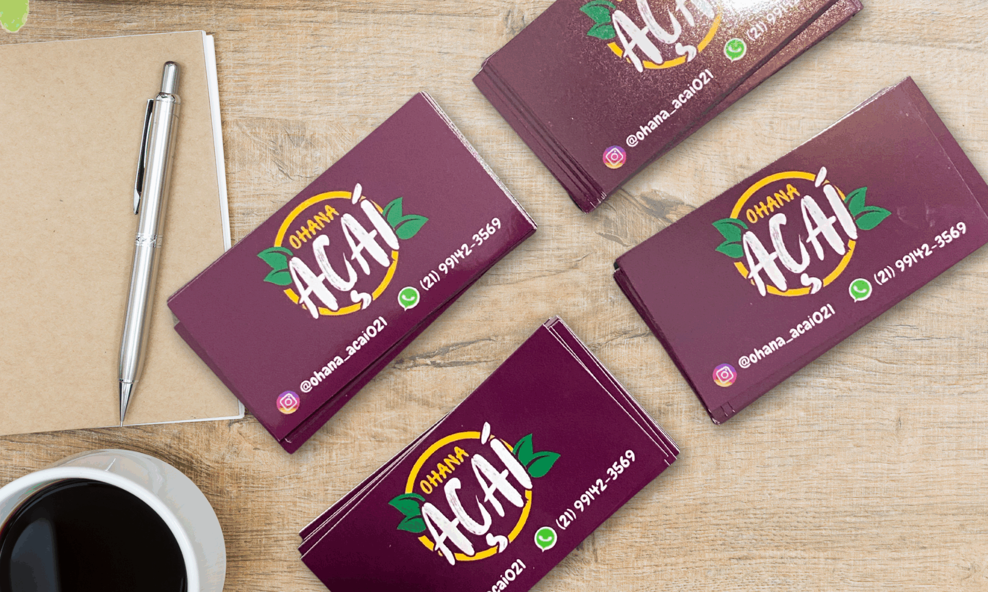 Cartões de visita personalizados com acabamento brilhante, exibindo o logotipo "Ohana Açaí", posicionados sobre uma mesa de madeira com itens de escritório. Imagem ideal para divulgar serviços de gráfica online, impressão de cartões de visita e materiais promocionais personalizados em pequenas e grandes quantidades.